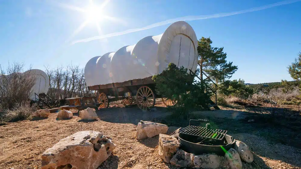 Covered wagons parked in a sunny, rocky landscape with a fire pit nearby.